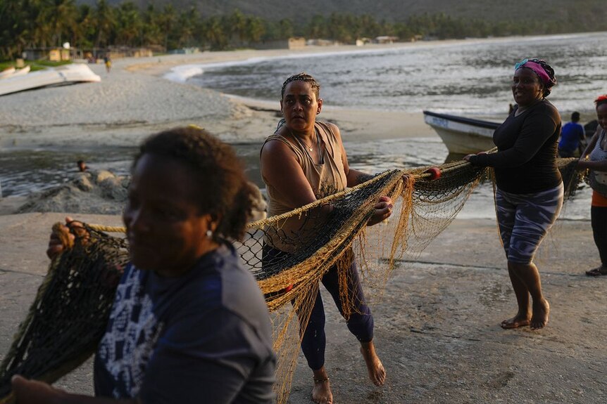 A group of women carry a fishing net back to the beach, their boat can be seen in the background.