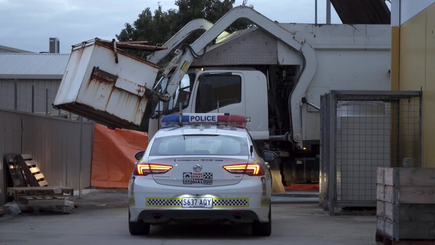 A police car in front of a truck holding a skip bin