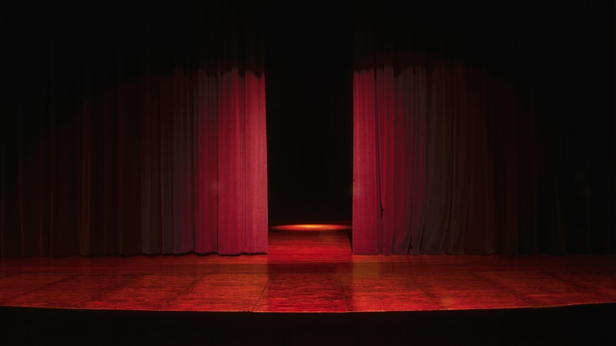 A dimly lit stage in a theatre with a row of chairs down the front. The red curtains are parted to show some of the stage.