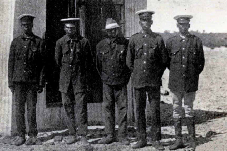 Five officers in dark uniforms stand in front of a corrugated iron shed.