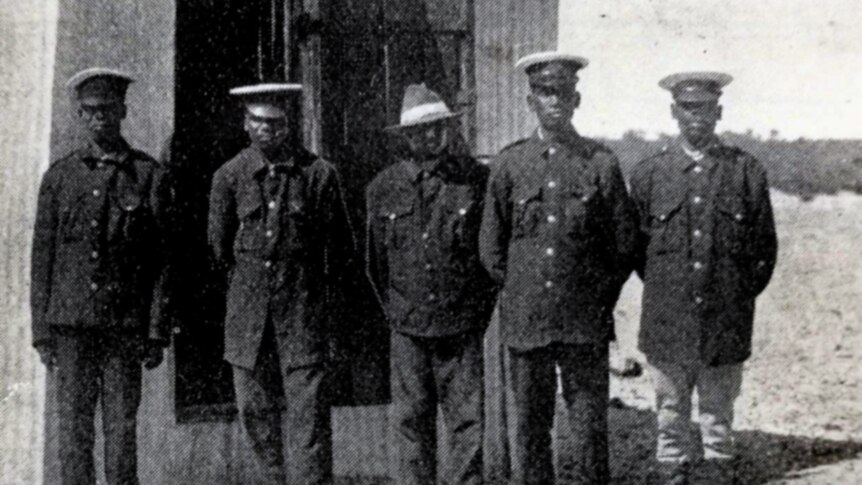 Five officers in dark uniforms stand in front of a corrugated iron shed.