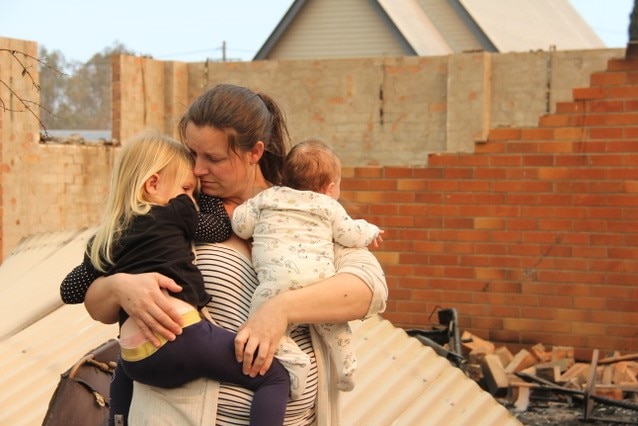 A woman holds her two children in front of a destroyed home.