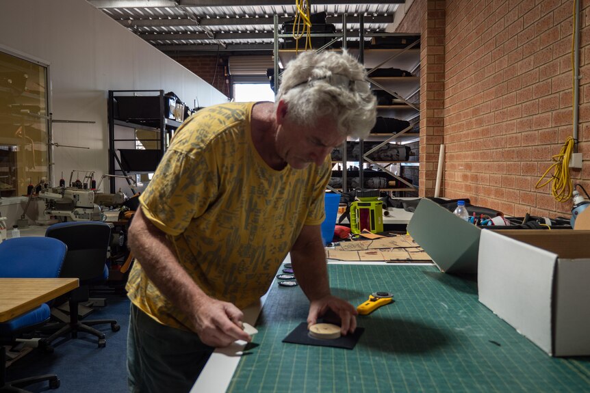 A grey-haired man in a yellow t-shirt leans over a worktable doing work. 