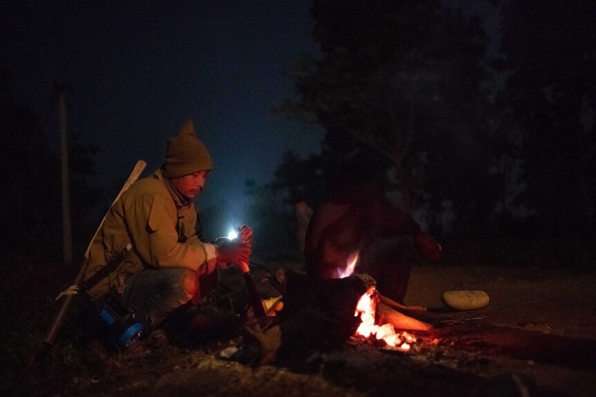 A farmer sits by an open fire at night.