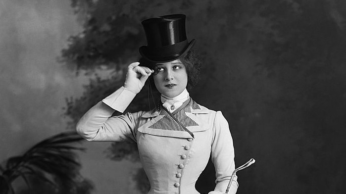 An early photograph shows Edna May in top hat while wearing a full-length hourglass-shaped coat, and posing with a monocle.