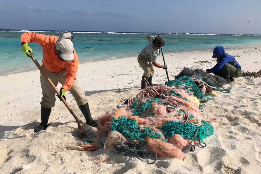 Three people are seen on a beach with a blue sky behind them and clear waters as they work at a long line of fishing nets.