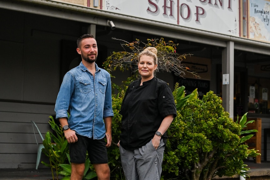 A man and a woman standing in front of a cafe and looking at the camera. 