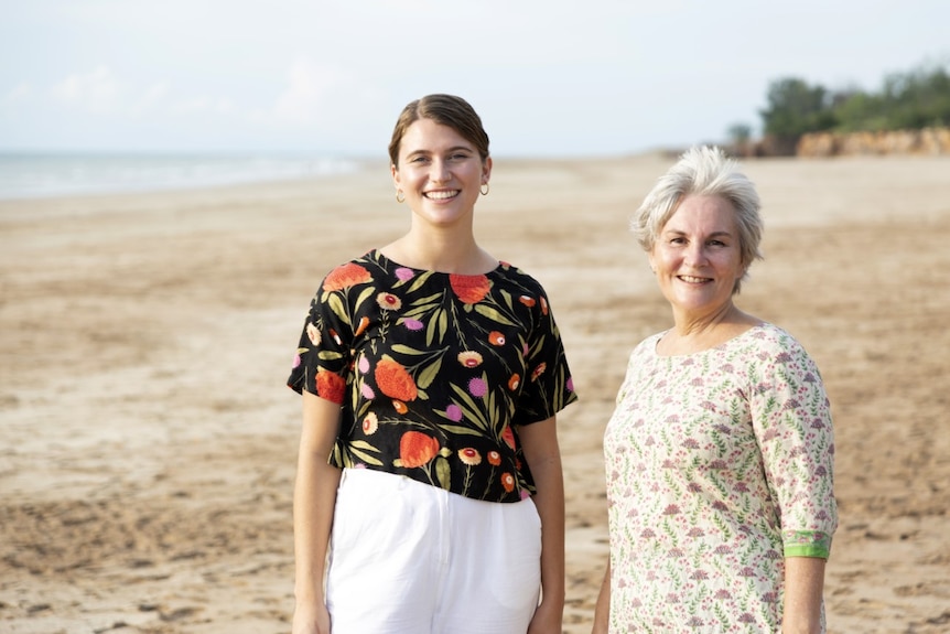 Two smiling women standing on a sandy beach. One is taller and younger, the other has short grey hair