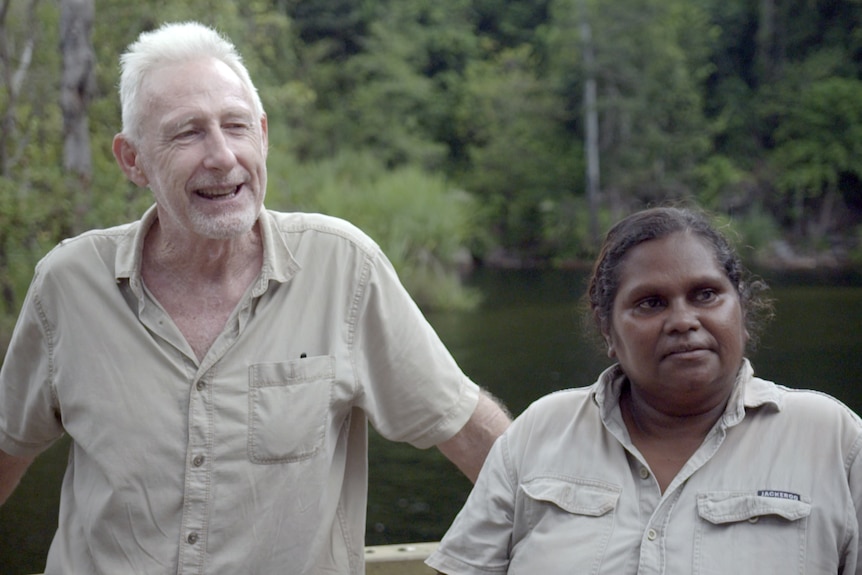 A man and woman, both in khaki shirts, stand by a railing with a waterfall behind them.