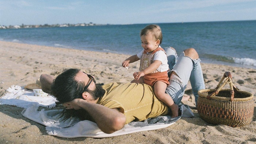 Man and his toddler daughter sitting on him while on the beach for a story about where new dads can find support.