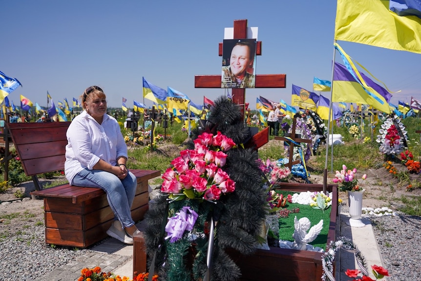 A woman who is upset looks on, while sitting on a bench next to a grave