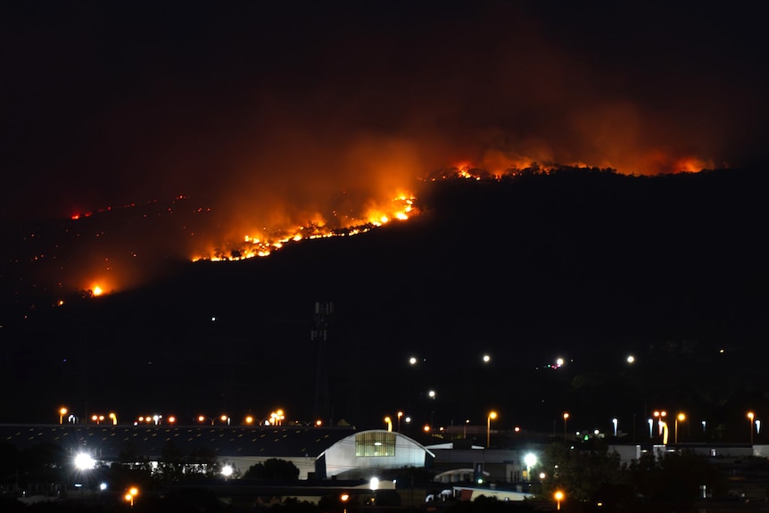 Bright flames from a bushfire in Perth's hills visible at night from far away, with buildings and lights in the foreground.