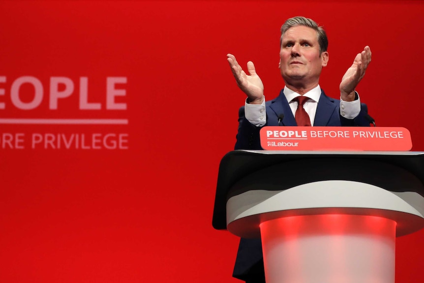 A man in a suit is speaking behind a lectern. The backdrop behind him is bright red.