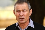 A man wearing a black jumper and collared shirt looks head on at the camera.