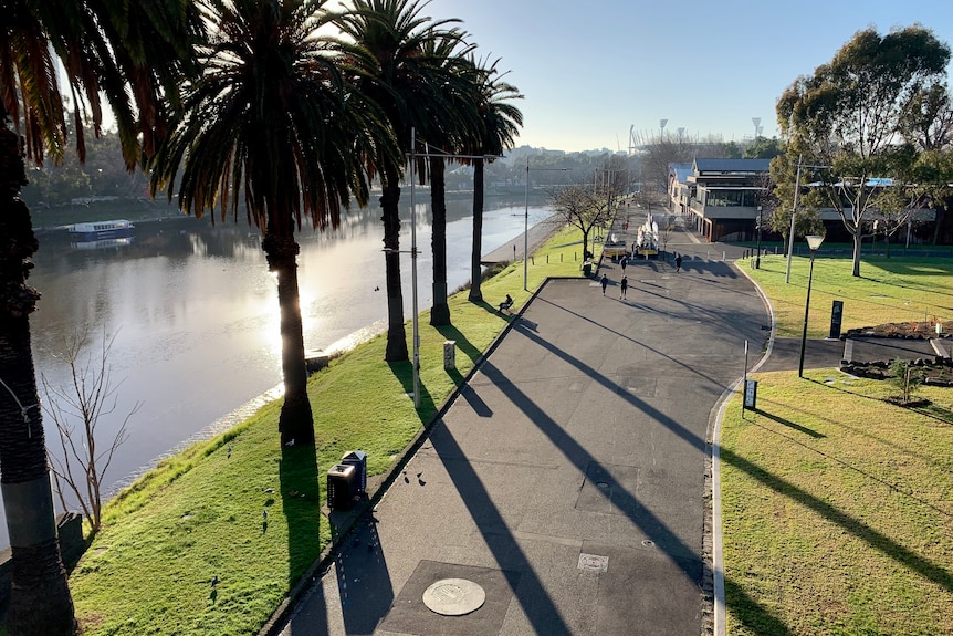 An elevated view of the footpath alongside the Yarra River, where just two or three people are walking in winter sunlight.