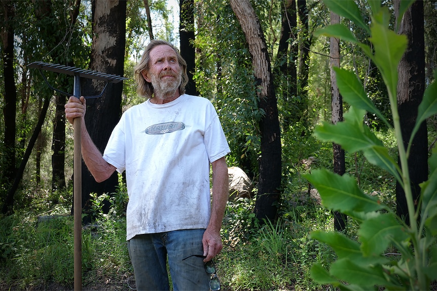 An older, bearded man in a white shirt stands among some trees, holding a rake.
