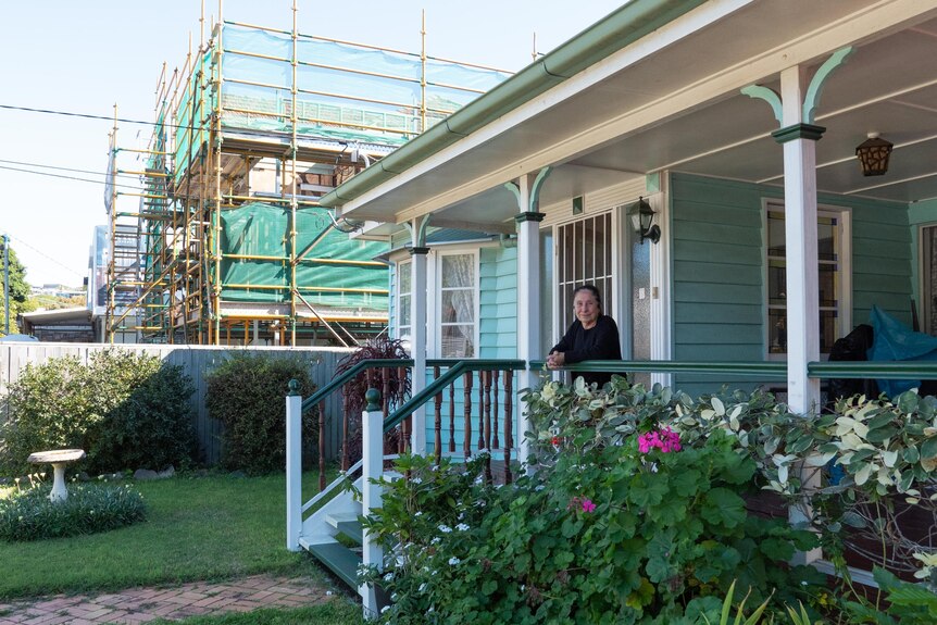A woman stands on her front deck. In the background her neighbour's house it covered in scaffolding.