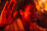 A close-up of woman of colour under red lighting, screaming and pressing her hands against glass.