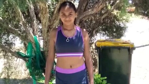 Denishar wears shorts and a crop top, smiling, in front of a tree with hose in the background.