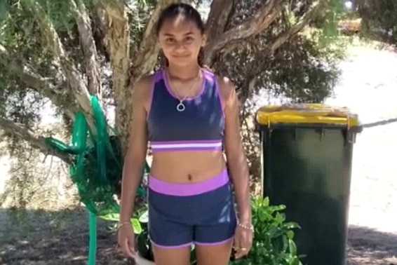 Denishar wears shorts and a crop top, smiling, in front of a tree with hose in the background.