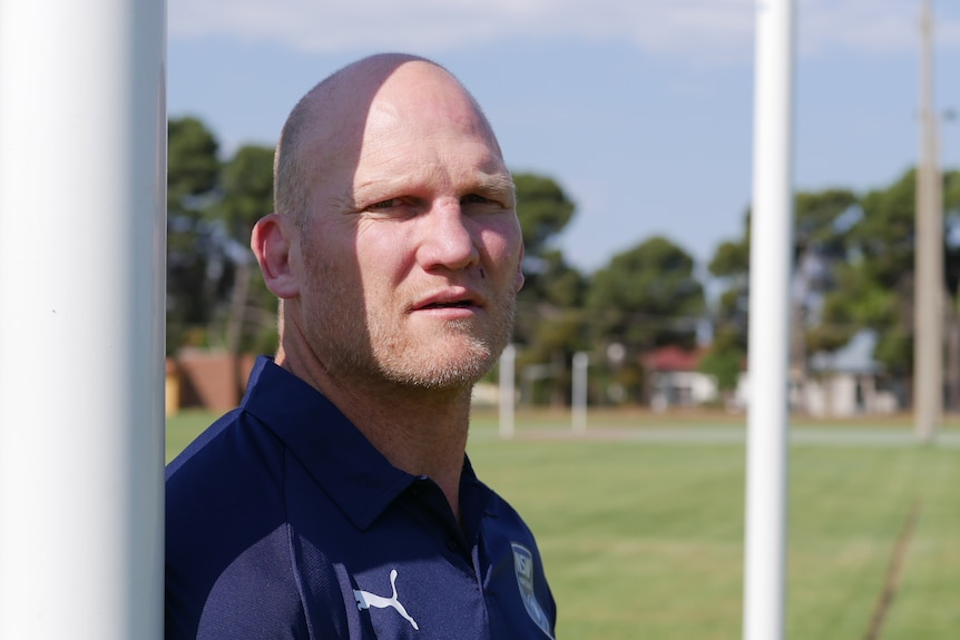 A middle-aged man with a bald head looks at the camera while leaning against a goal post on a rugby field.
