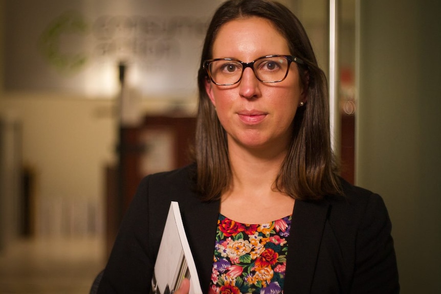 A woman with brown hair and glasses wearing a black jacket and floral top looks at the camera.