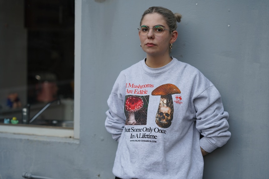 A woman in her mid 20s with glasses and hair up in buns stands in front of a grey wall, she's wearing a graphic sweater