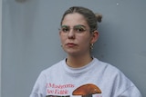 A woman in her mid 20s with glasses and hair up in buns stands in front of a grey wall, she's wearing a graphic sweater