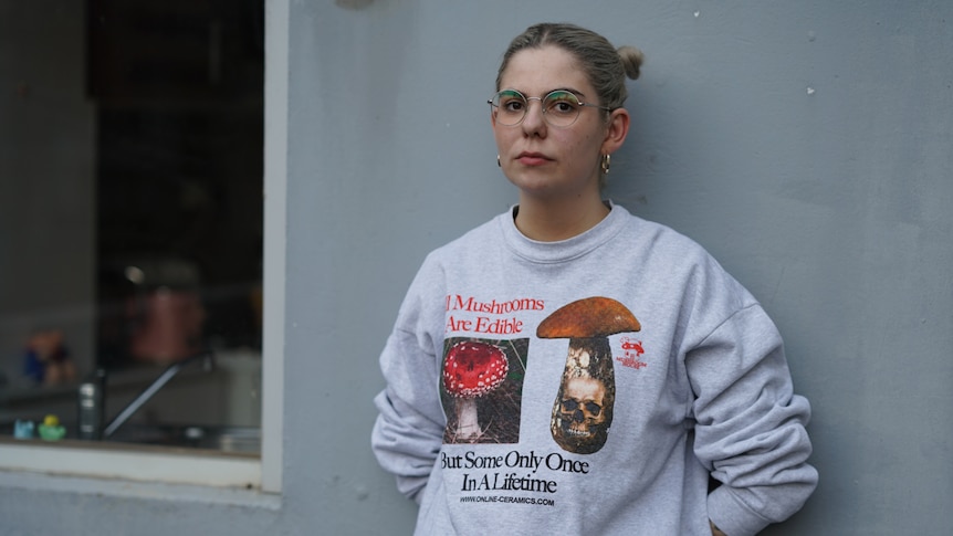 A woman in her mid 20s with glasses and hair up in buns stands in front of a grey wall, she's wearing a graphic sweater