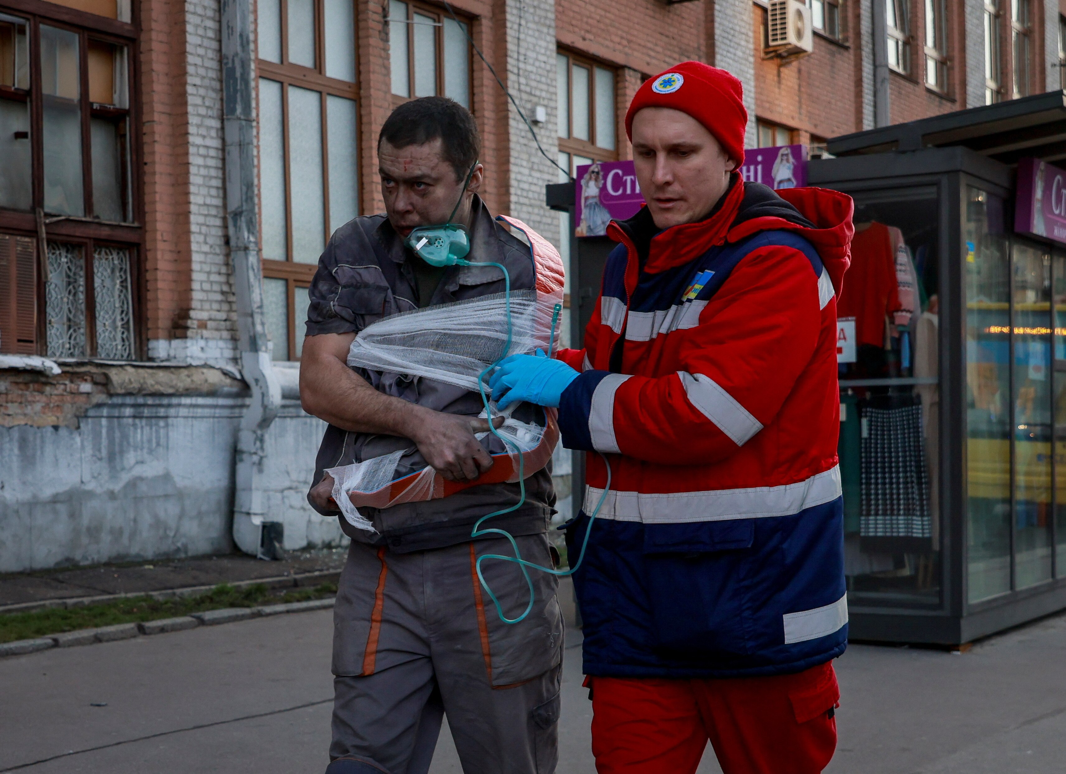 Two men walking next to a building. One is a medical worker, who holds the arm of the other man, whose arm is bandaged.