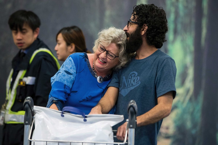 A smiling woman leans on an also smiling man while pushing a luggage trolley.