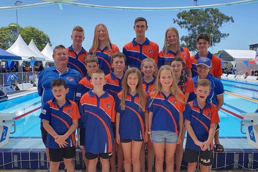 A group of people of varied ages smiling and standing in a cluster wearing orange and blue shirts in front of a swimming pool.