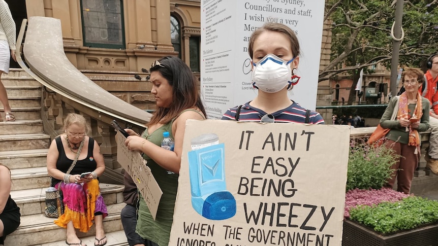 Girl in face mask with a sign that says 'It ain't easy being wheezy when the govt ignores climate change...'