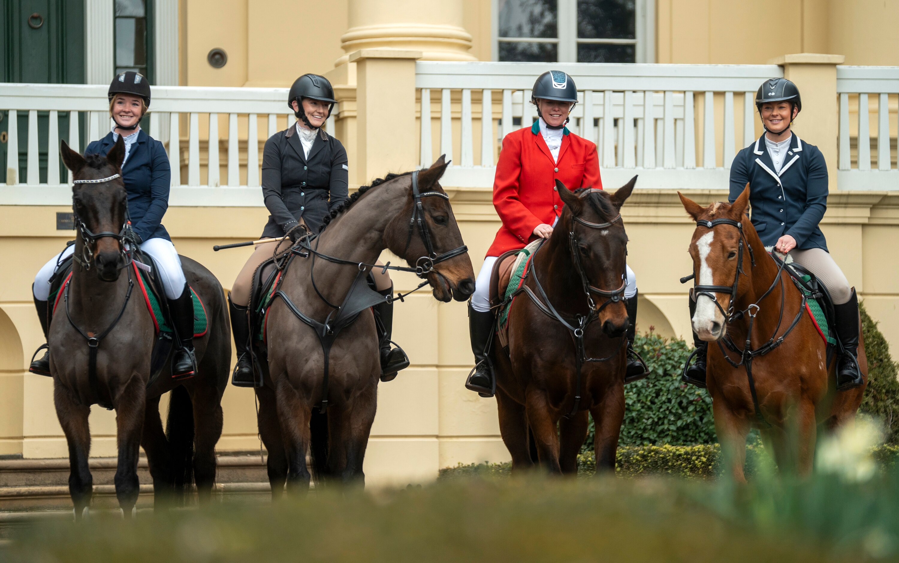Four women wearing navy, red and black blazers pose for a photo on their horses outside a daffodil-coloured mansion.