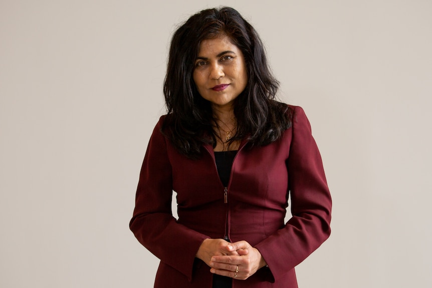 A woman with long black hair wearing a maroon suit jacket and black top stands upright with her hands folded.