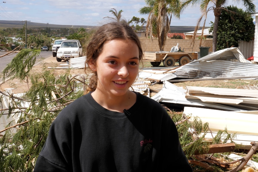 A young girl wearing a black jumper stands in front of a street with debris and a tree on the ground after a cyclone.