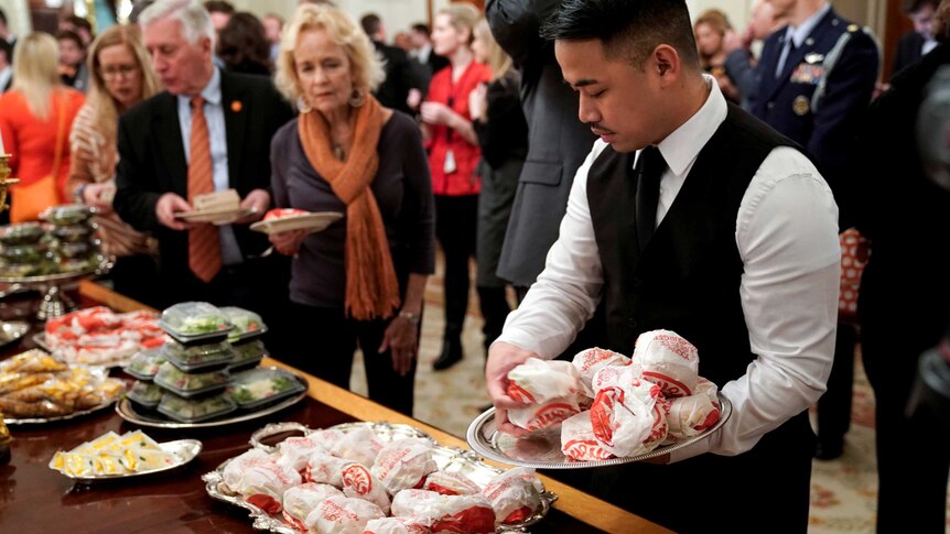A White House server places Wendy's burgers onto a tray while crowds of people gather around the table.