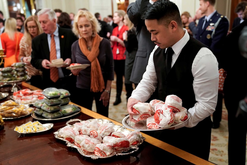 A White House server places Wendy's burgers onto a tray while crowds of people gather around the table.