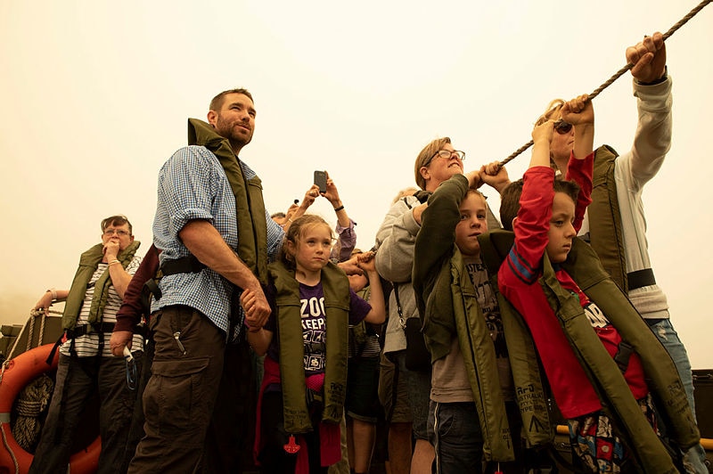 Children and a handful of adults wear life jackets as several of them hold onto a rope for balance. The sky is hazy.