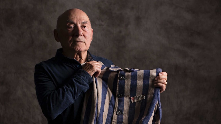 An older man sits on a stool while holding up a striped jacket, dappled grey background.