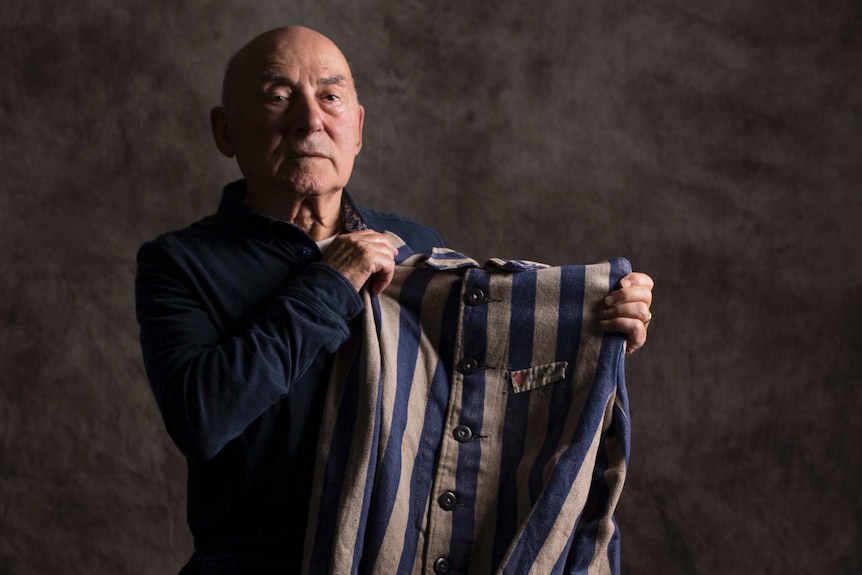 An older man sits on a stool while holding up a striped jacket, dappled grey background.