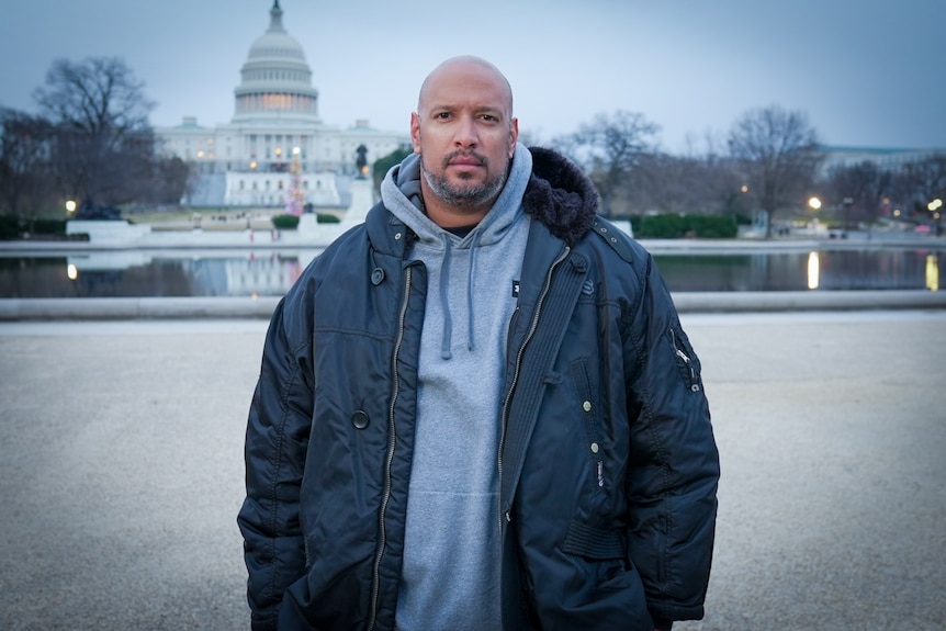 An African American man in a dark coat and hoodie stands outside the US Capitol building