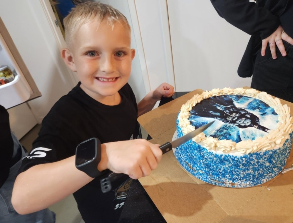 Un niño pequeño de cabello rubio sonríe mientras sostiene un cuchillo sobre un pastel.