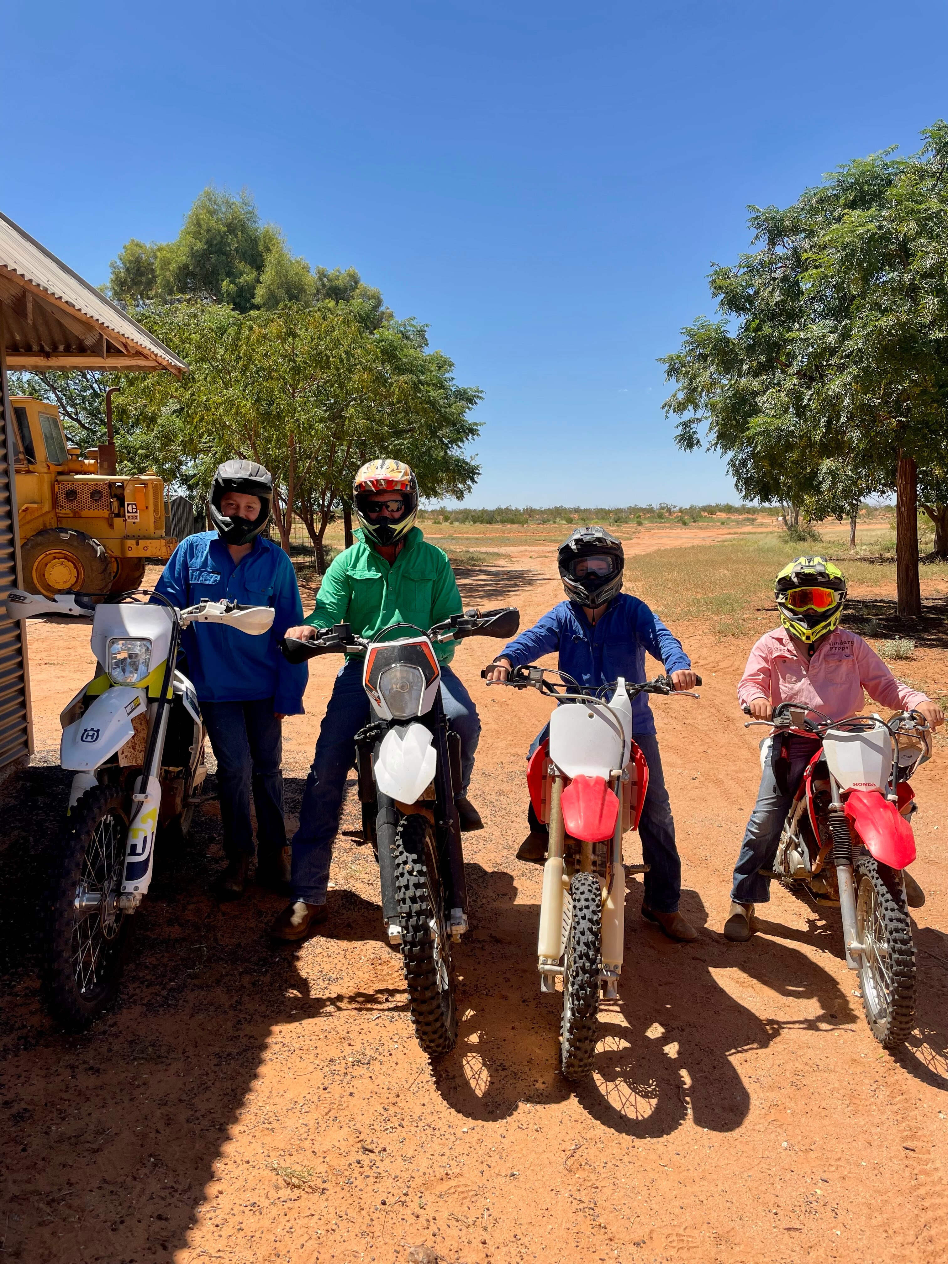 Quatro pessoas em motocicletas, com uma estrada de terra ao fundo.