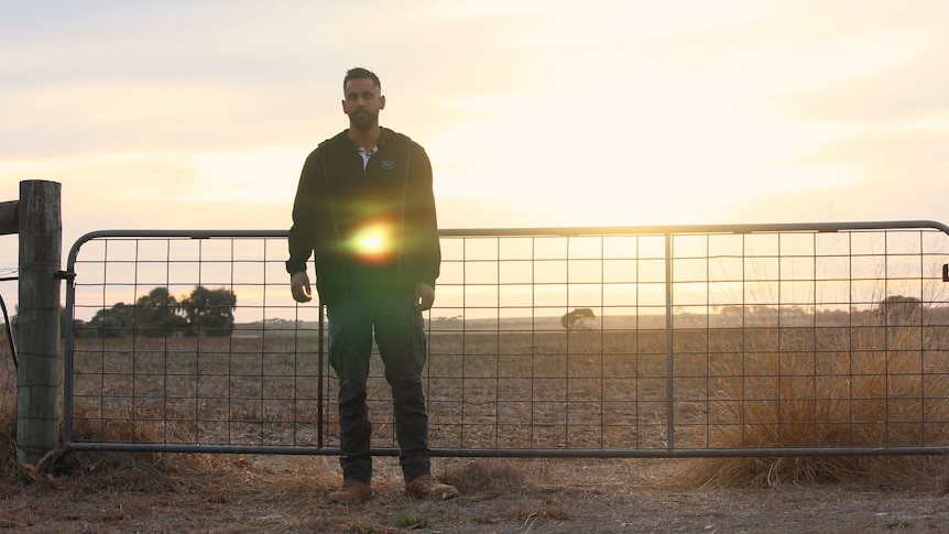 A man stands in front of a gate at sunrise with the sun shining behind him.