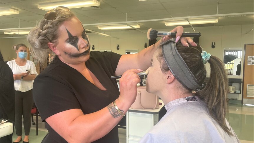 A student in creepy clown makeup is applying makeup to another student's face. 