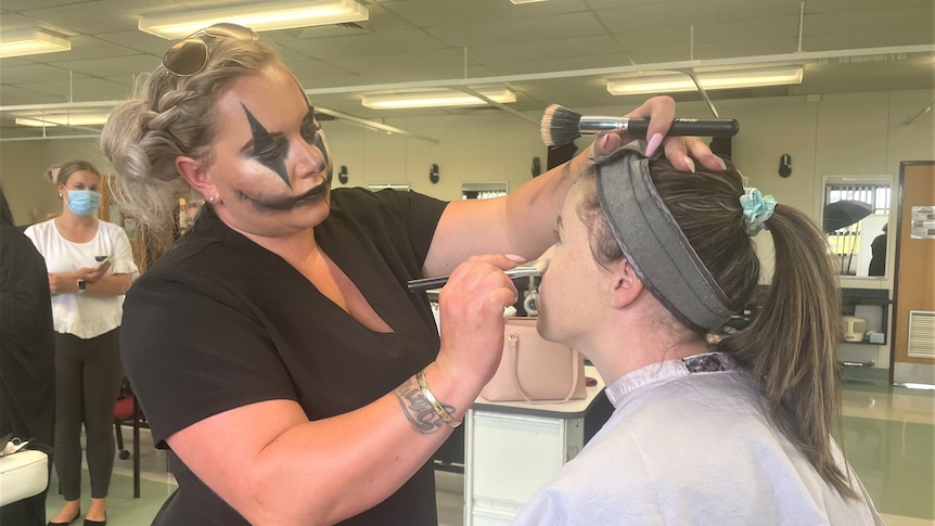A student in creepy clown makeup is applying makeup to another student's face. 