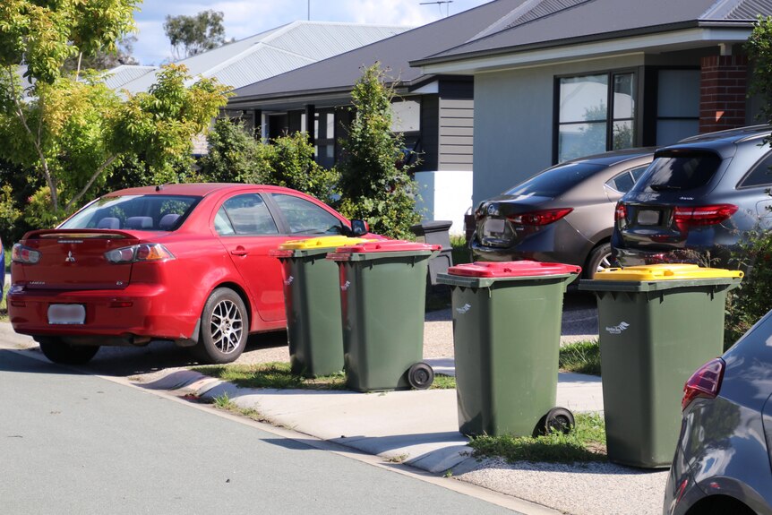Two red top and two yellow top council bins on the curb with four cars parked around them