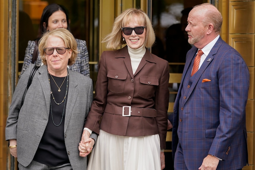 A blonde woman with large dark glasses smiling and holding hand ofher female lawyer as she leaves court