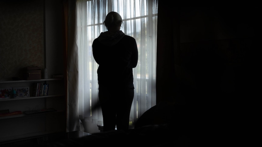 A woman stands in front of a window with a sheer curtain, in shadow 